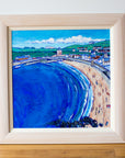A coastal painting of Eyemouth Harbour in a limewaxed chuncky wood frame. It sits on a wood floor and is leaning against a white wall. 