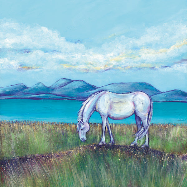 A white pony stands grazing on a grassy bank in front of calming turquoise waters.Hills can be seen in the background against a light blue sky with a smattering of white clouds with yellow undertones. 