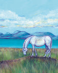 A white pony stands grazing on a grassy bank in front of calming turquoise waters.Hills can be seen in the background against a light blue sky with a smattering of white clouds with yellow undertones. 