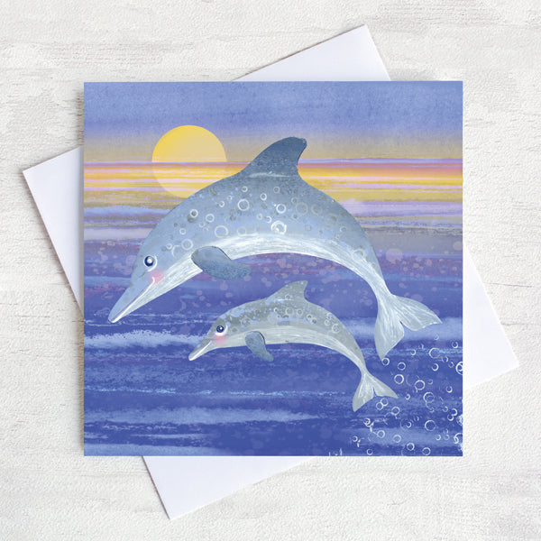 A nostalgic seaside greetings card designed by Joanne Wishart, featuring a leaping dolphin and her young splashing out of the water in front of an evening sunset.