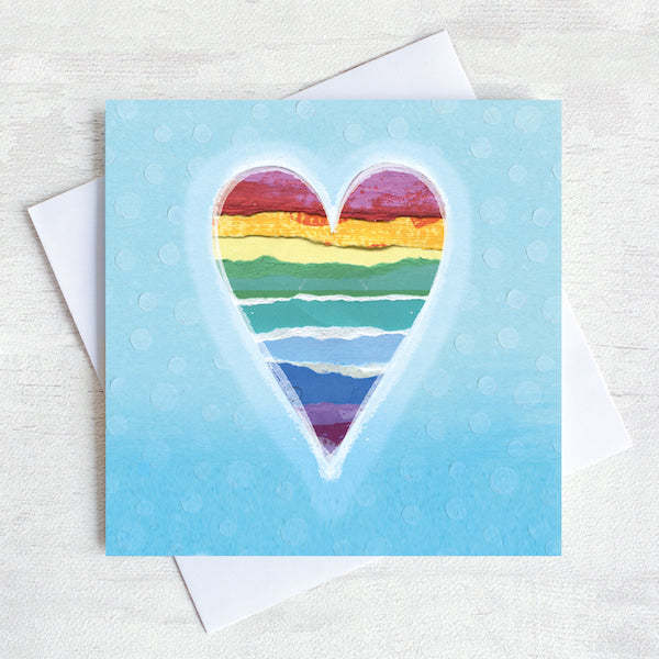 A Valentines Greetings Card featuring a rainbow striped heart  on a place blue background.