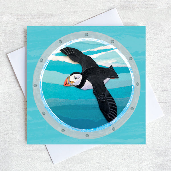 A greetings card featuring a flying puffin as viewed through a ships porthole.