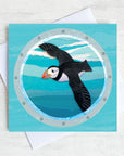 A greetings card featuring a flying puffin as viewed through a ships porthole.