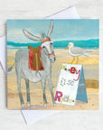 A coastal greetings card featuring a seaside donkey chatting to a seagull who is sitting on top of a sign advertising Donkey rides.