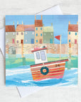 A charming coastal greetings card featuring a paper collage of a fishing boat in front of a row of colourful cottages in a harbour.