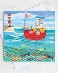 A whimsical greetings card of a fishing boat with a lighthouse  and a shoal of fish swimming in the sea.