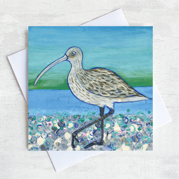 A greetings card featuring a curlew on a pebbled beach with a sea green background.