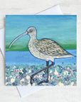 A greetings card featuring a curlew on a pebbled beach with a sea green background.