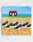 A greetings card featuring a flock of oystercatchers on a pebbled beach with a fisherman's hut in the background.