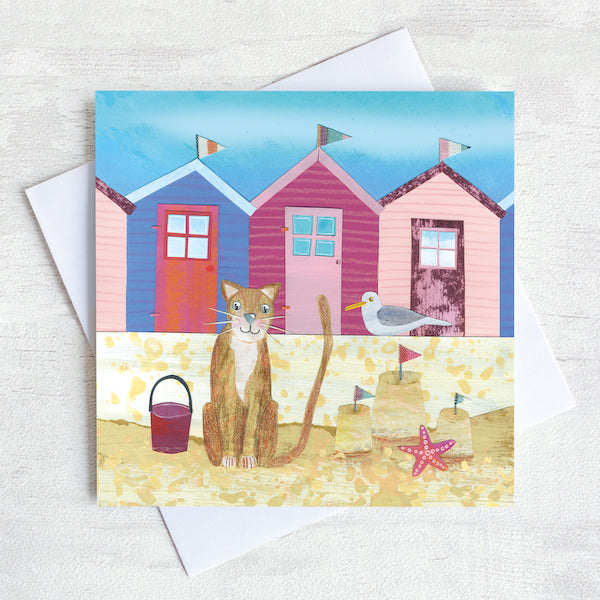 A seaside greetings card featuring a ginger cat on a beach infant of a row of colourful beach huts.