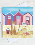 A seaside greetings card featuring a ginger cat on a beach infant of a row of colourful beach huts.