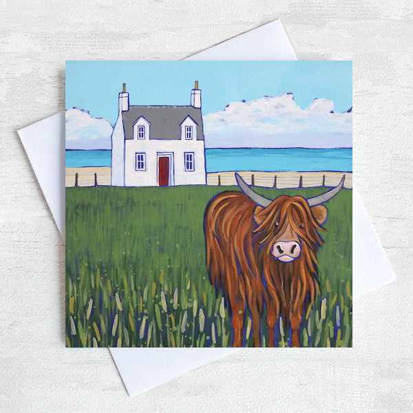 A greetings card featuring a highland cow and a white cottage with a turquoise sea in the distance. 