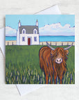 A greetings card featuring a highland cow and a white cottage with a turquoise sea in the distance. 