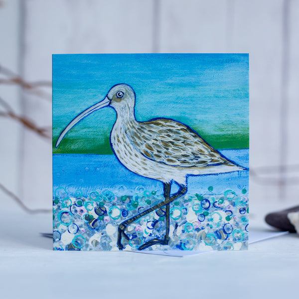 A greetings card featuring a curlew on a pebbled beach with a sea green background.