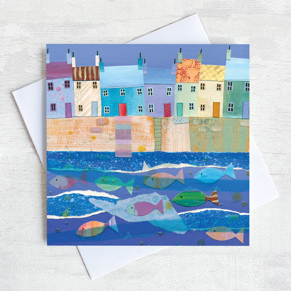 A quirky greetings card featuring a row of colourful cottages on a harbour wall with a shoal of colourful fish in the water.