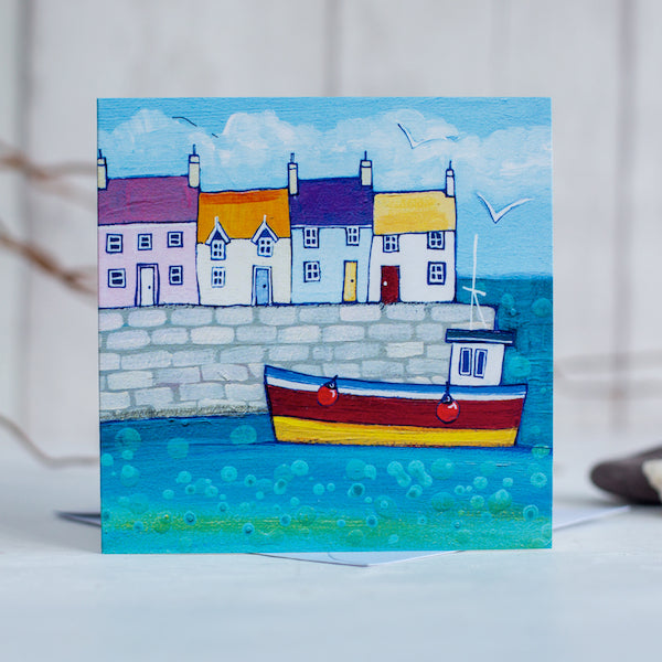 A coastal greetings card featuring a cute harbour with colourful cottages and a fishing boat bobbing on the water.