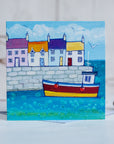 A coastal greetings card featuring a cute harbour with colourful cottages and a fishing boat bobbing on the water.
