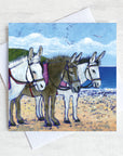 A greetings card featuring three fluffy seaside donkeys on a beach.