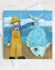 A greetings card featuring a fisherman in yellow dungarees holding up a giant fish!