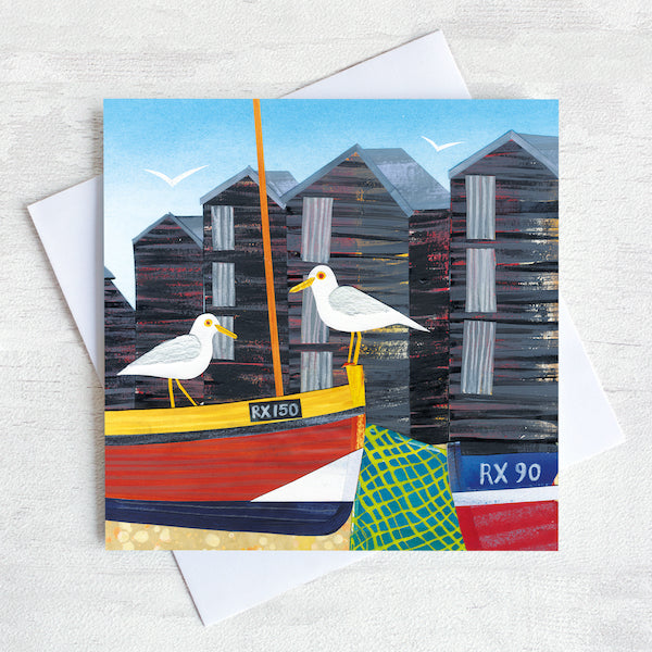 A charming seaside greetings card featuring a cute little harbour with seagulls perched on the cable boats infant of the net sheds.