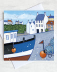 A greetings card featuring a painting of Crail on the Fife coastline. There is a fishing boat in the foreground that is surrounded by creels and lobster pots.