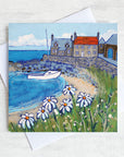A greetings card featuring a colourful painting of craster harbour with daises in the foreground and a sailing boat in the bay.