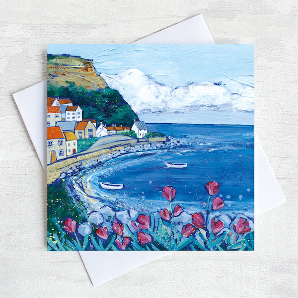 A greetings card featuring a painting of Brunswick bay with pink flowers in the foreground and a blue sky above the quaint village on the cliff side.