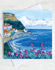 A greetings card featuring a painting of Brunswick bay with pink flowers in the foreground and a blue sky above the quaint village on the cliff side.