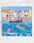 A greetings card of coastal harbour featuring seals bobbing in the water around a fishing boat.