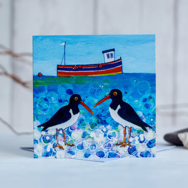 A charming greetings card featuring oystercatchers chatting on a pebbled beach with a cute little fishing boat bobbing on a teal green sea.