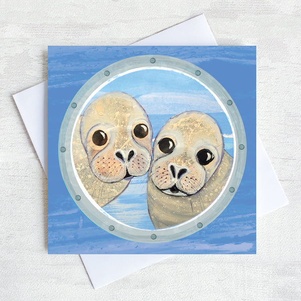 A greetings card featuring two cheeky seals peeking through a ships porthole.