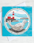 A greetings card featuring a grey seal basking on the rocks in front of a red and white lighthouse.