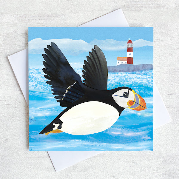 A coastal greetings card with a flying puffin swooping over a quirky red and white lighthouse.