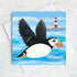 A coastal greetings card with a flying puffin swooping over a quirky red and white lighthouse.