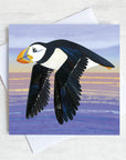 A quirky greetings card featuring a flying puffin gliding over the sea in the evening light. 