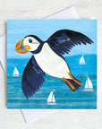 A coastal greetings card featuring a flying puffin over a turquoise sea. 