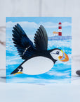 A greetings card featuring a flying puffin in front of a quirky little lighthouse.