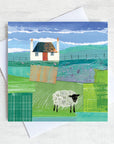 A scottish greetings card featuring a collaged artwork of a traditional blockhouse with a red door and a black headed sheep grazing in the field in front of the house.