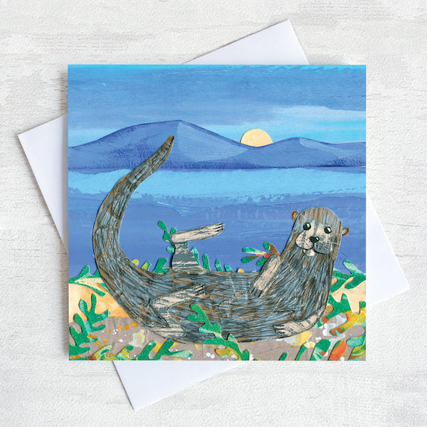 A greetings card with a sea otter curled on a moonlit beach surrounded by seaweed. 
