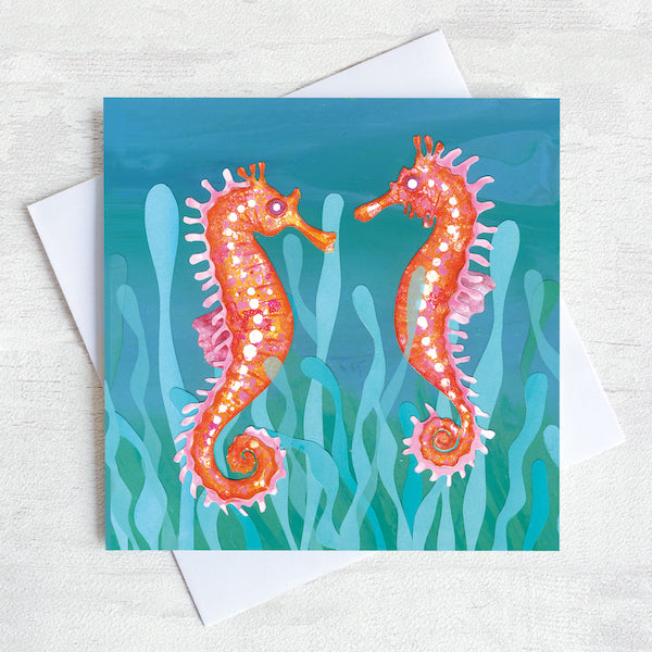 A teal green greetings card featuring two orange sea horses bobbing in the seaweed.