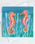 A teal green greetings card featuring two orange sea horses bobbing in the seaweed.