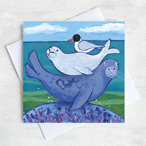 A coastal greetings card featuring a seal bathing on a rock with a baby on it&#39;s back and back headed gull. 