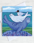 A coastal greetings card featuring a seal bathing on a rock with a baby on it's back and back headed gull. 