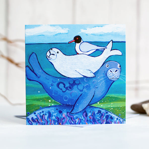 A greetings card featuring a cute family of seals snuggled on each others back, with a black headed gull on top.