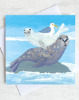 A cutsy seaside greetings card featuring a seal with it's pup on its back with a friendly seagull.