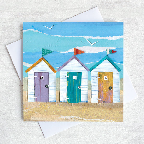 A colourful coastal greetings card featuring 3 beach huts all in a row on a golden sand beach.