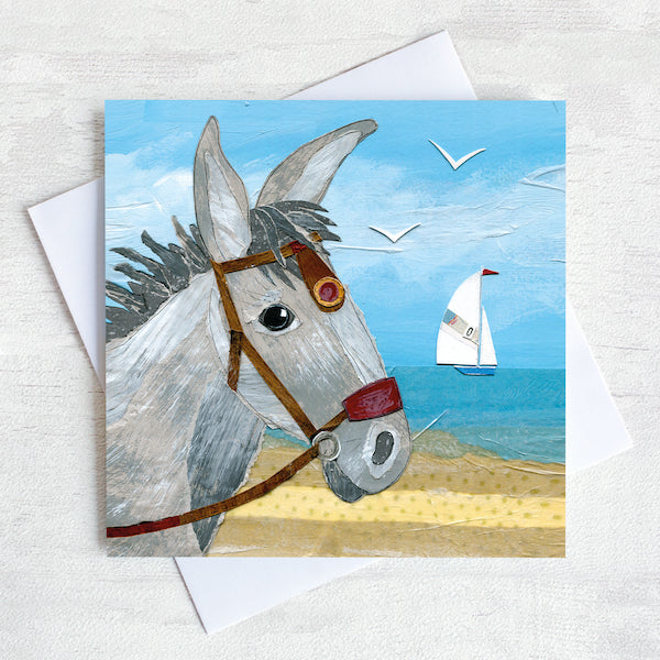 A coastal greetings card featuring the portrait head of a seaside donkey standing on a beach with a sailing boat in the distance on the sea.