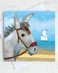 A coastal greetings card featuring the portrait head of a seaside donkey standing on a beach with a sailing boat in the distance on the sea.