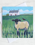 A scottish greetings card featuring a white stone cottage with a black headed sheep in the field in front with a starling on its back.