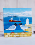 A greetings card featuring two quirky black headed gulls infront of a view to the sea and the farne islands with a bright red fishing boat on the water.